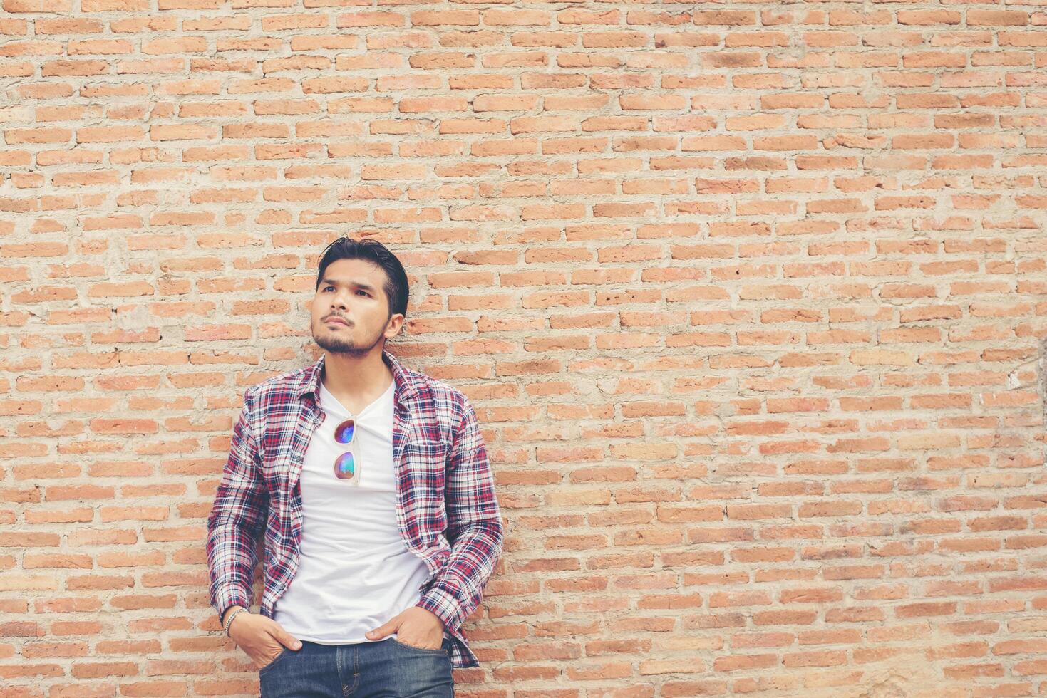 joven apuesto hombre hipster con camisa a cuadros y jeans contra una pared de ladrillos. foto