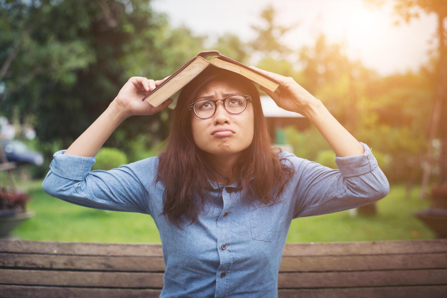 Young hipster woman cover her head with book, afraid of something falling while sitting on bench in the park. photo