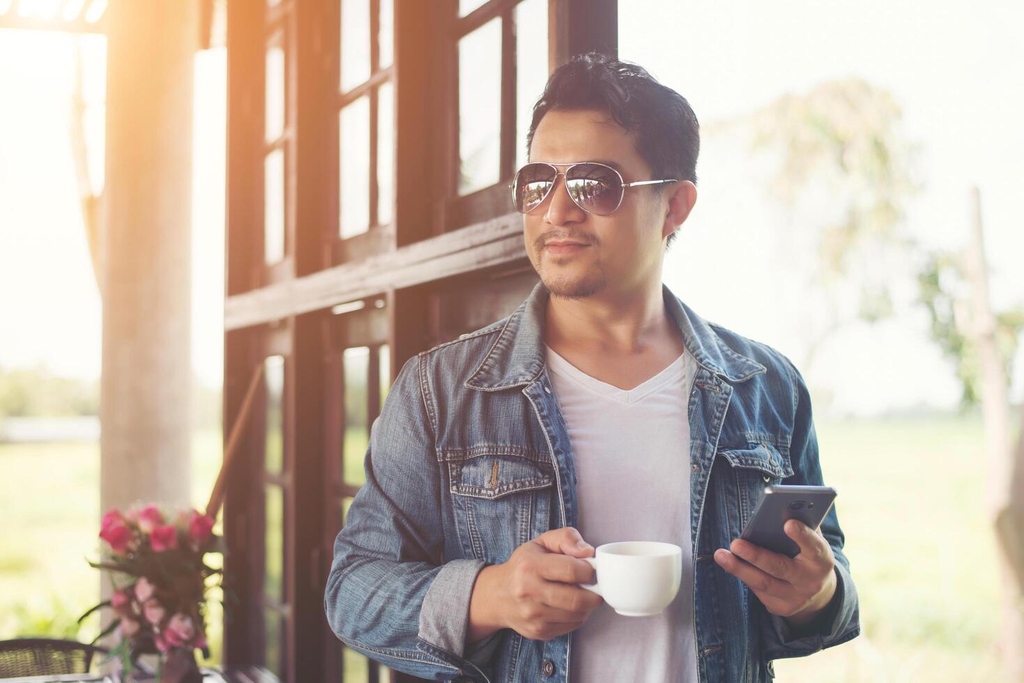 hombre hipster sosteniendo una taza de café y caminando en la cafetería, relajando el teléfono mirando hacia otro lado. foto