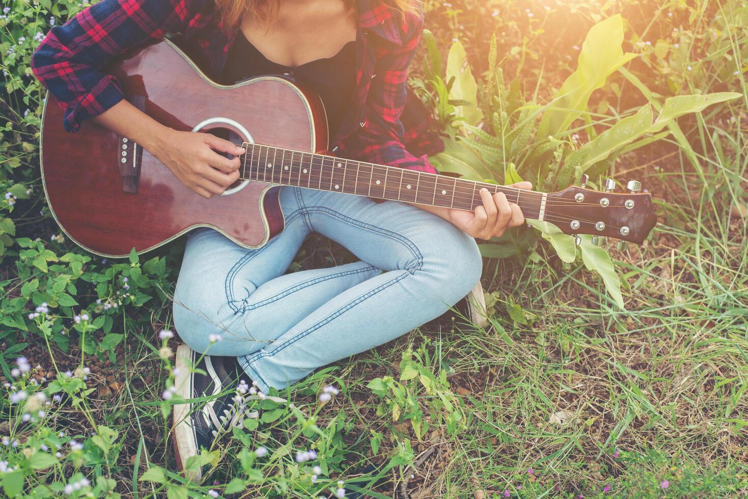 manos de mujer hipster tocando la guitarra sentada en el césped en el parque, relajándose con la naturaleza. foto