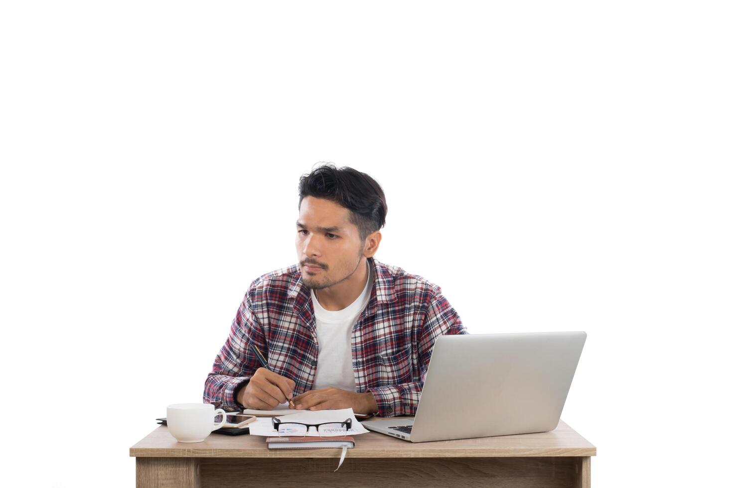 Thoughtful young man  looking away while writing sitting at his working place isolated on white background. photo