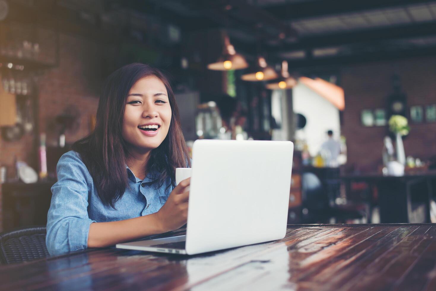 Beautiful young hipster woman sitting in a coffee shop, holding a cup of coffee relax and play with her laptop, looking away, happy and fun. Lifestyle concept. photo