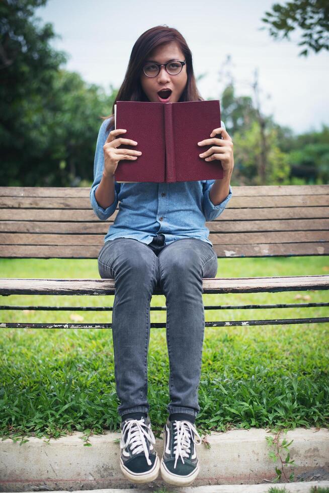 Charming hipster girl was surprised, reading a novel while sitting on bench in the park. photo