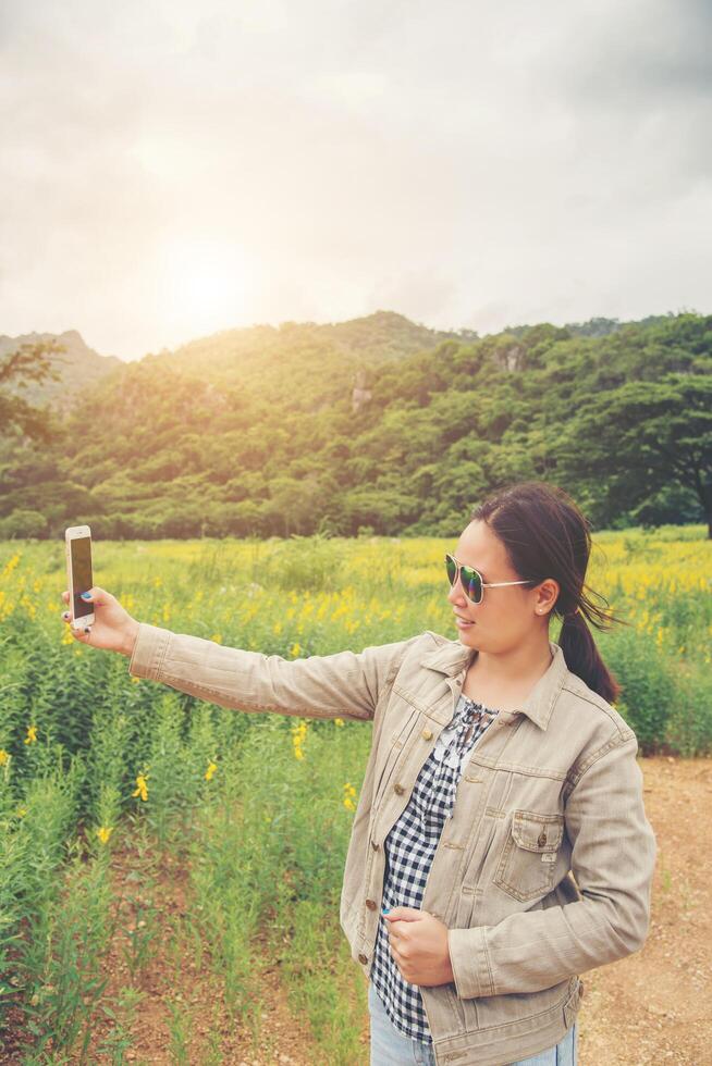 Closeup of beautiful teenage girl in yellow flower field taking a selfie on smartphone outdoors in summer. photo