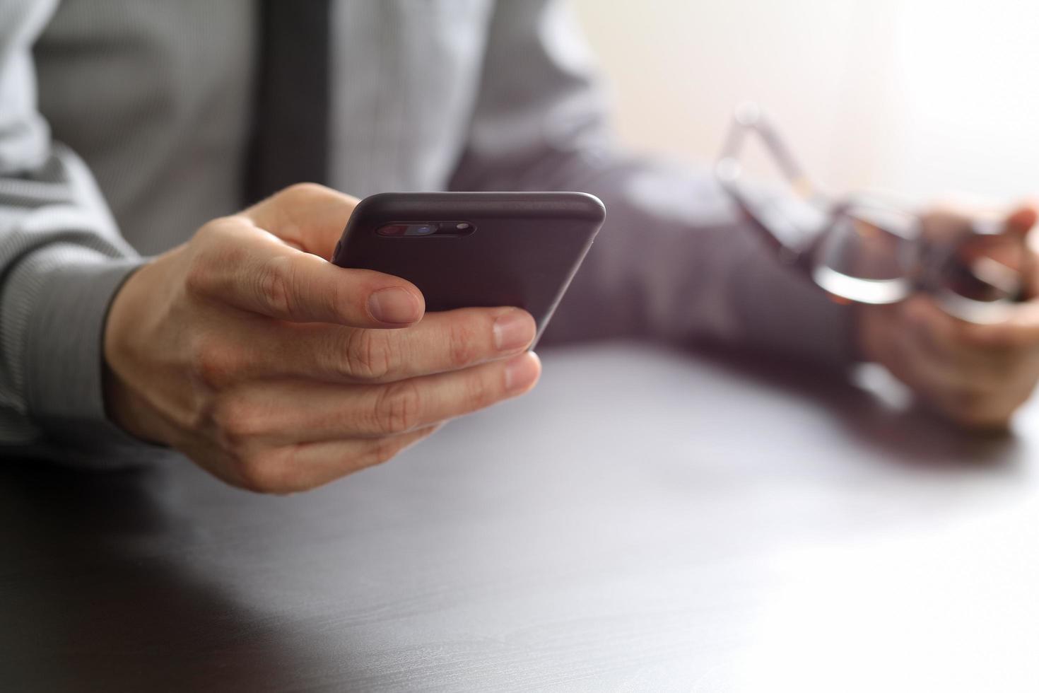 close up of businessman working with mobile phone and eyeglass on wooden desk in modern office photo