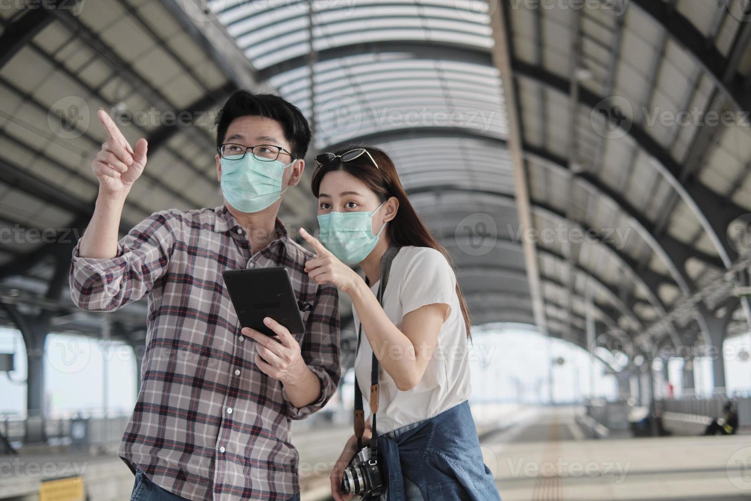 Thai man with face mask helps Asian female tourist search information, find travel locations with tablet at train station platforms, passenger trip lifestyles, casual journey vacation transportation. photo