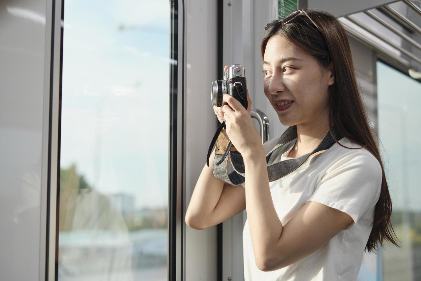 A Beautiful Asian female tourist with camera in the passenger cabin, traveling by sky train, taking snapshot photos when transporting in urban view, city lifestyle by railway, happy journey vacation.