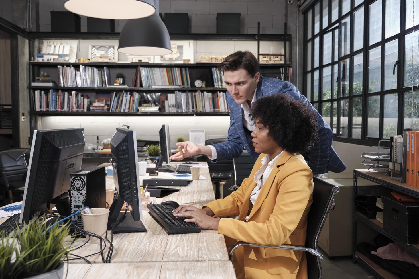 Young African American female worker sits, works with computer, brainstorming, talking, and discussing with Caucasian male colleague and partnership about business jobs in workspace office company. photo