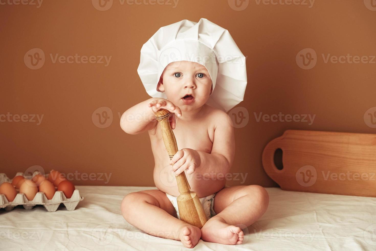 Little baby boy in a chef's hat sitting on the table holds a wooden rolling pin on a brown background photo