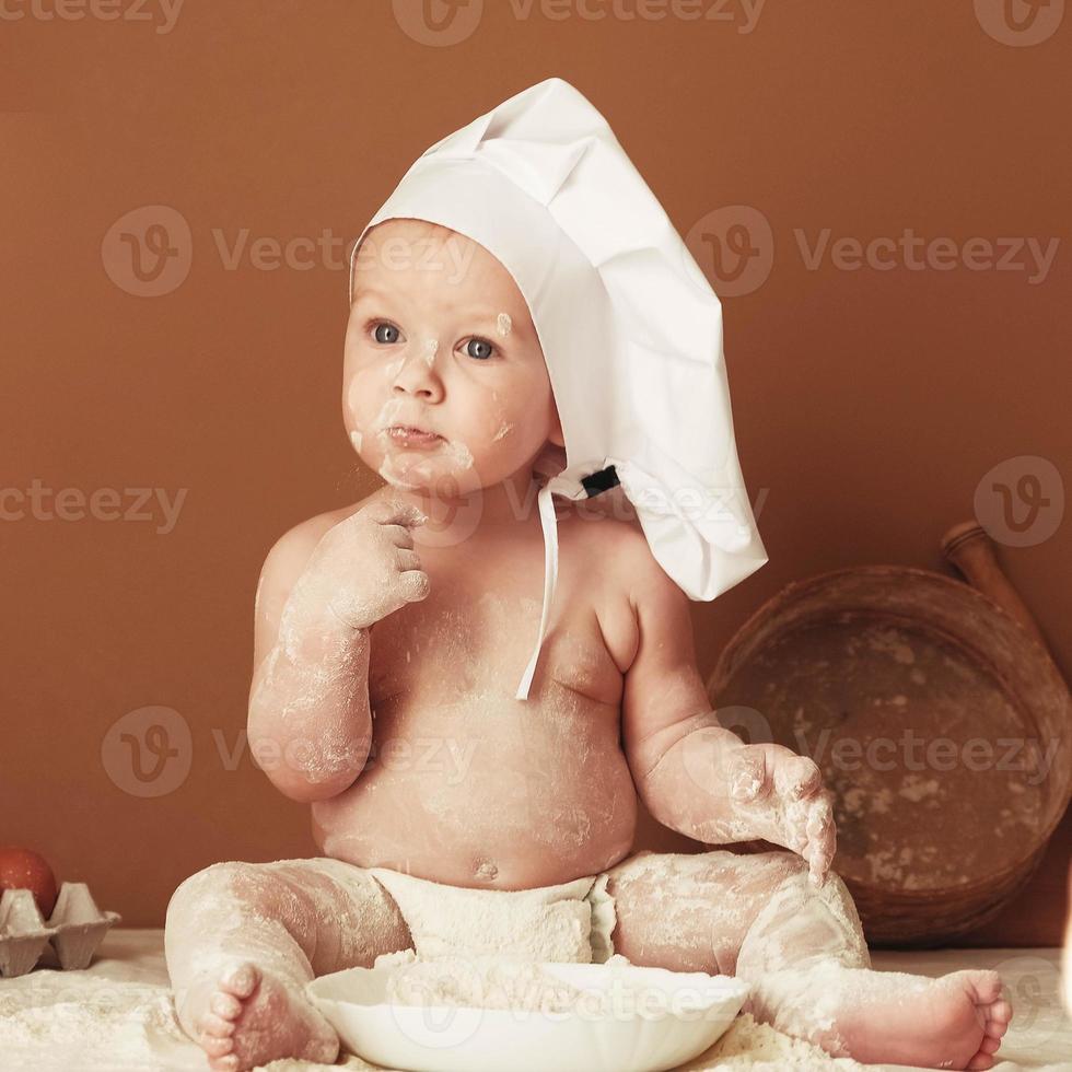 Little boy baker in a chef's hat sitting on the table playing with flour on a brown background with a wooden rolling pin, a round rustic sieve and eggs. Copy, empty space for text photo