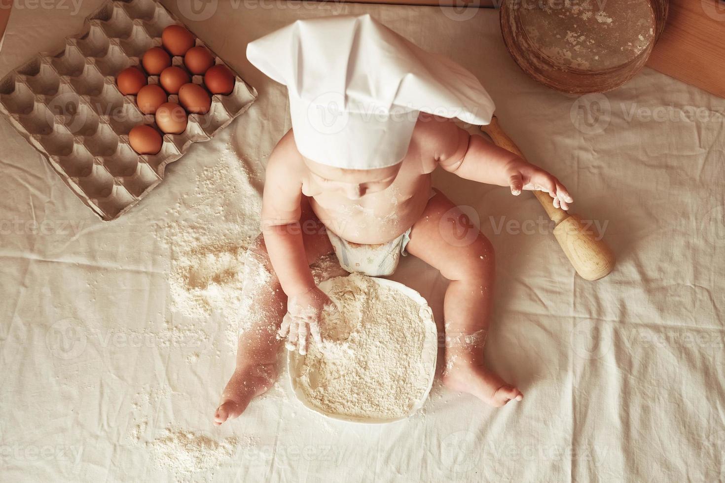 Little boy baker in a chef's hat sitting on the table playing with flour on a brown background with a wooden rolling pin, a round rustic sieve and eggs. Top view. Copy, empty space for text photo