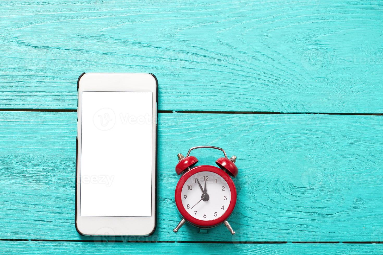 Mobile phone and red retro alarm clock with five minutes to twelve o'clock on blue wooden background. Top view and blank screen photo