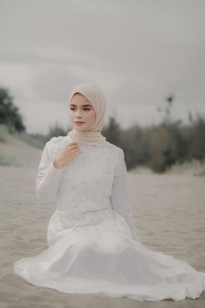 hermosa modelo femenina islámica usando moda hijab, un vestido de novia moderno para mujer musulmana sentada en la arena y la playa. retrata a una modelo asiática usando hiyab divirtiéndose en la playa con árboles foto