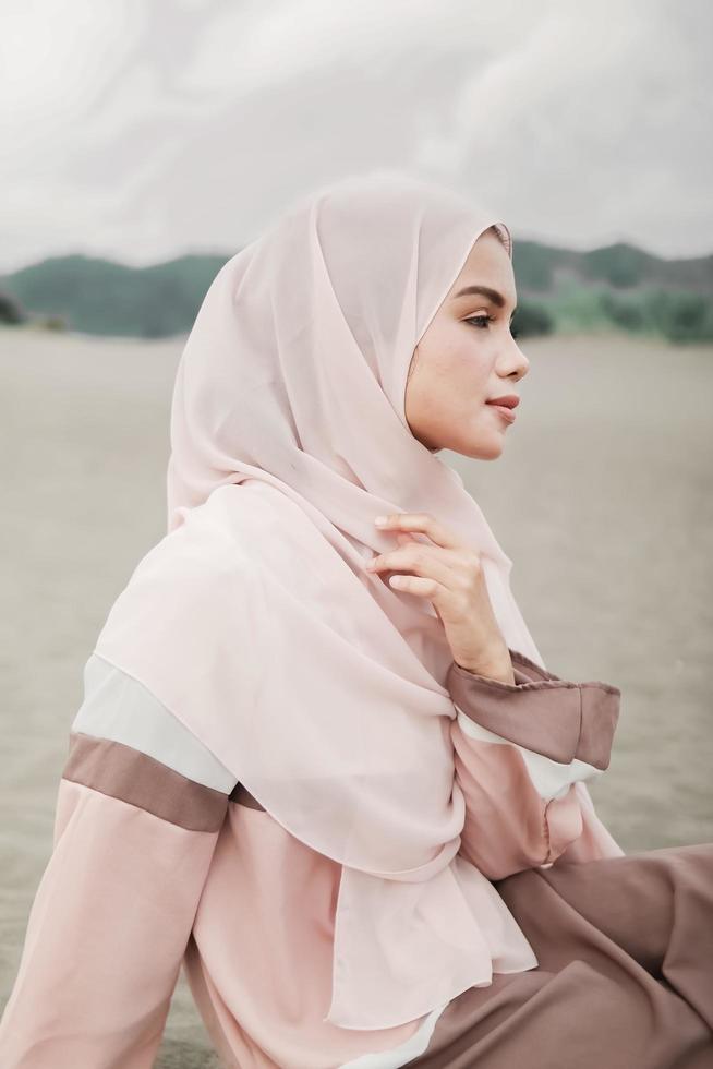 hermosa modelo femenina islámica usando moda hijab, un vestido de novia moderno para mujer musulmana sentada en la arena y la playa. retrata a una modelo asiática usando hiyab divirtiéndose en la playa con árboles foto