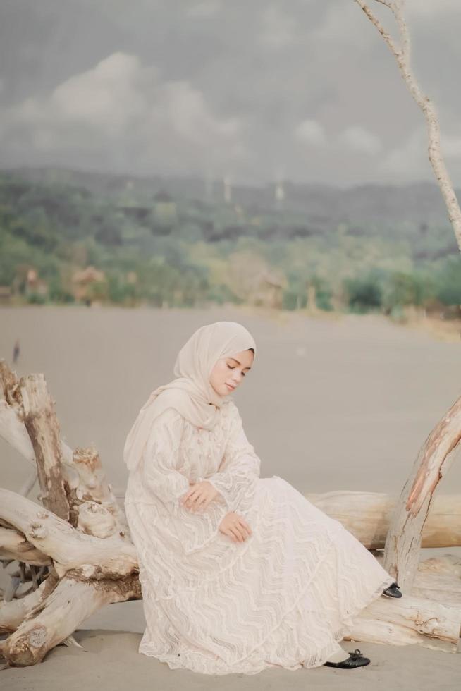 Beautiful islamic female model wearing hijab fashion, a modern wedding dress for muslim woman sitting in the sand and beach. Portrait an asian girl model using hijab having fun at the beach with trees photo