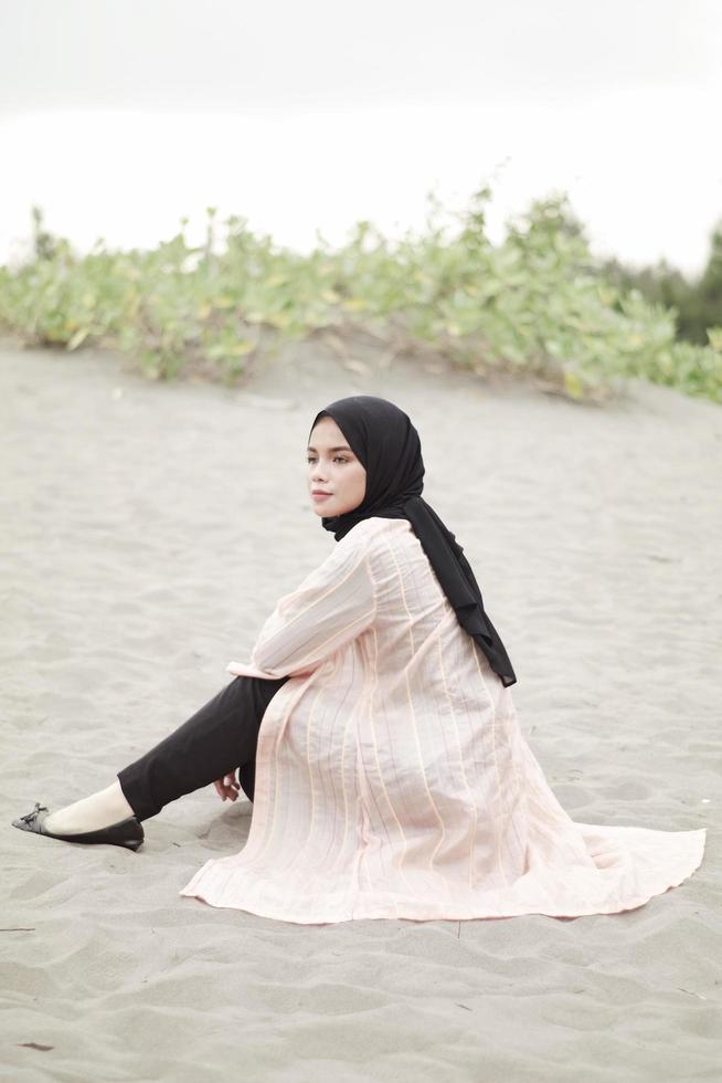 Beautiful islamic female model wearing hijab fashion, a modern wedding dress for muslim woman sitting in the sand and beach. Portrait an asian girl model using hijab having fun at the beach with trees photo