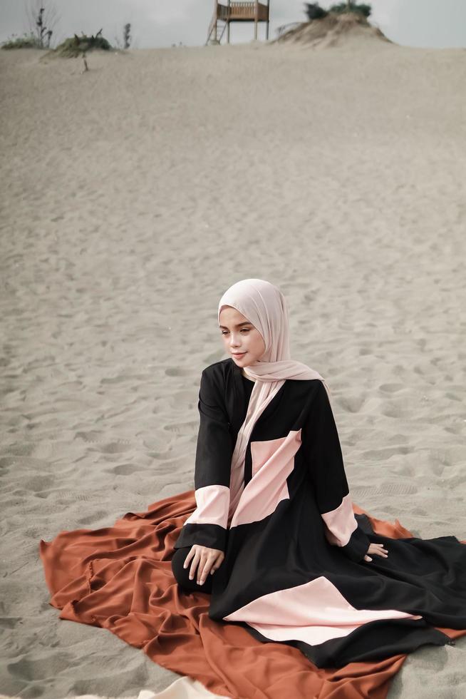 Beautiful islamic female model wearing hijab fashion, a modern wedding dress for muslim woman sitting in the sand and beach. Portrait an asian girl model using hijab having fun at the beach with trees photo