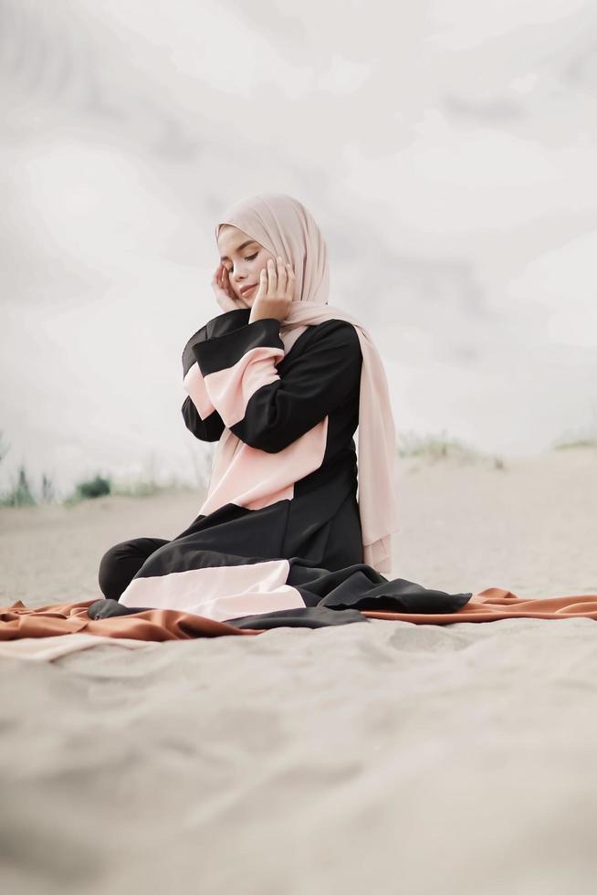 hermosa modelo femenina islámica usando moda hijab, un vestido de novia moderno para mujer musulmana sentada en la arena y la playa. retrata a una modelo asiática usando hiyab divirtiéndose en la playa con árboles foto