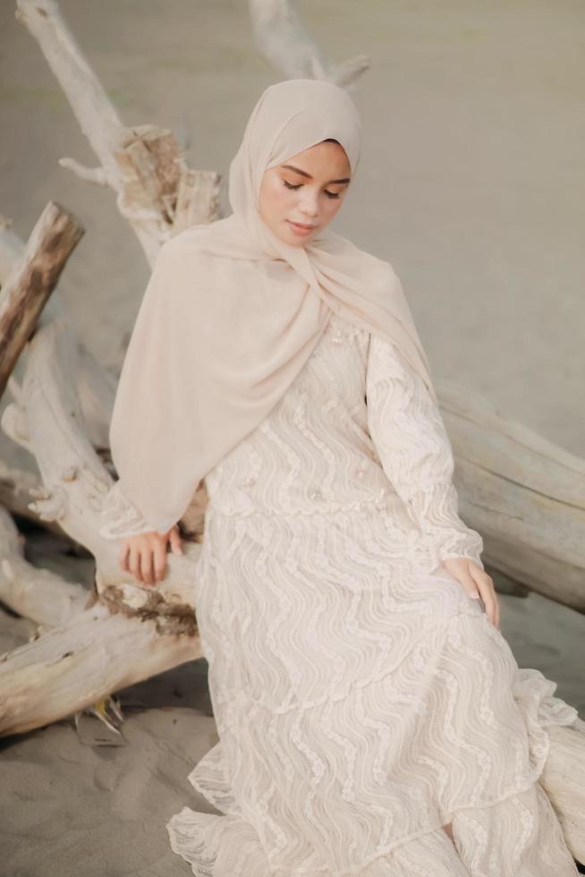 hermosa modelo femenina islámica usando moda hijab, un vestido de novia moderno para mujer musulmana sentada en la arena y la playa. retrata a una modelo asiática usando hiyab divirtiéndose en la playa con árboles foto