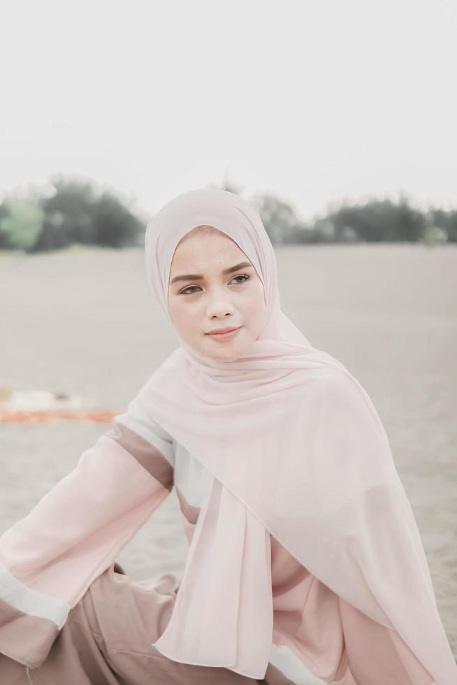 Beautiful islamic female model wearing hijab fashion, a modern wedding dress for muslim woman sitting in the sand and beach. Portrait an asian girl model using hijab having fun at the beach with trees photo