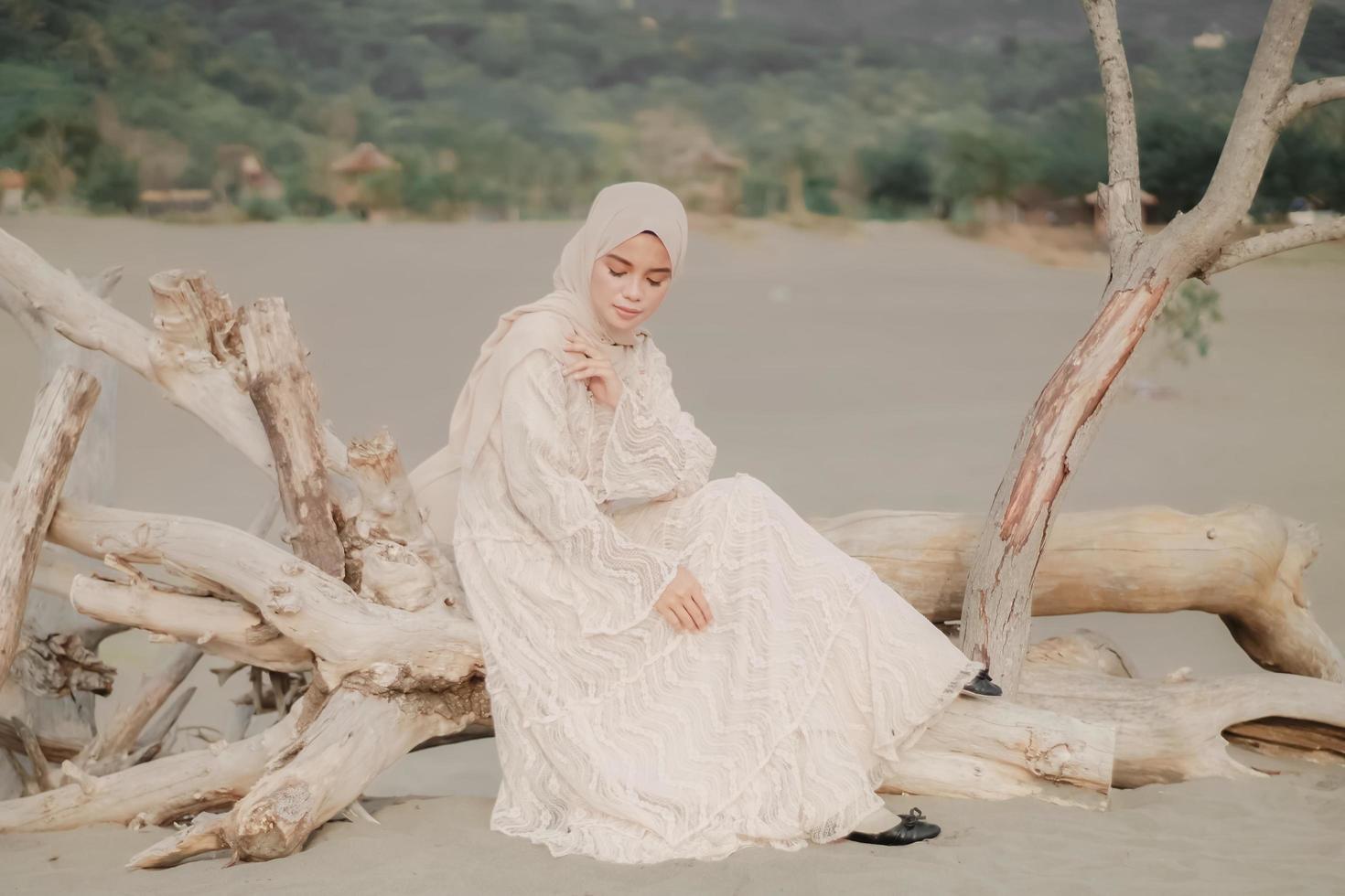 hermosa modelo femenina islámica usando moda hijab, un vestido de novia moderno para mujer musulmana sentada en la arena y la playa. retrata a una modelo asiática usando hiyab divirtiéndose en la playa con árboles foto