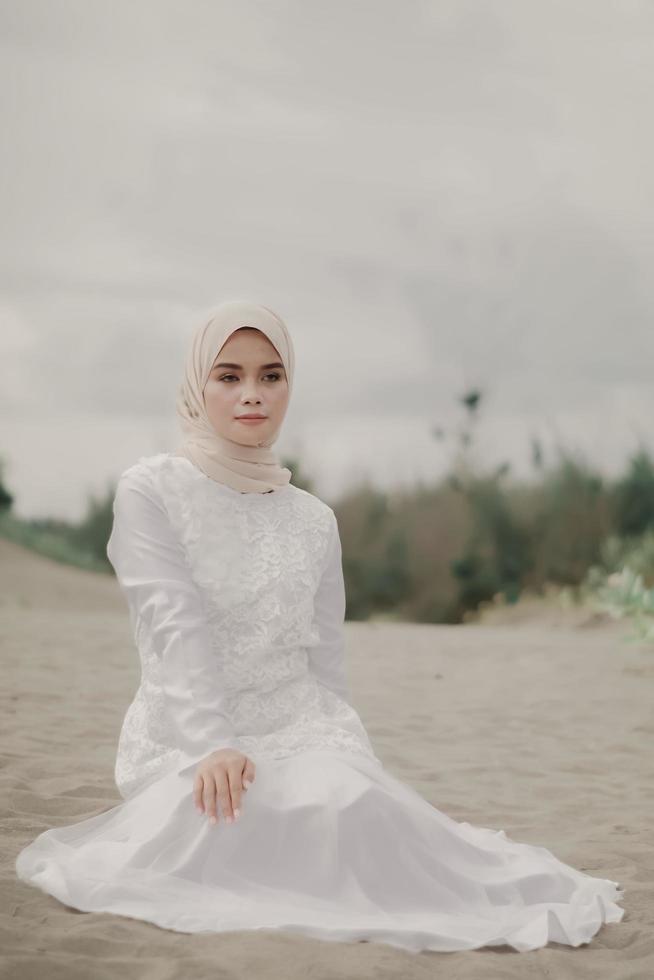 Beautiful islamic female model wearing hijab fashion, a modern wedding dress for muslim woman sitting in the sand and beach. Portrait an asian girl model using hijab having fun at the beach with trees photo