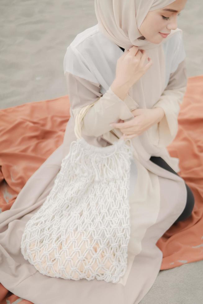 Beautiful islamic female model wearing hijab fashion, a modern wedding dress for muslim woman sitting in the sand and beach. Portrait an asian girl model using hijab having fun at the beach with trees photo