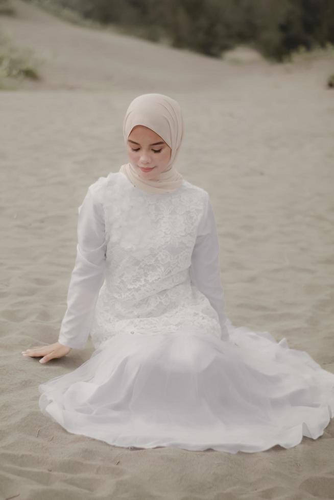 hermosa modelo femenina islámica usando moda hijab, un vestido de novia moderno para mujer musulmana sentada en la arena y la playa. retrata a una modelo asiática usando hiyab divirtiéndose en la playa con árboles foto