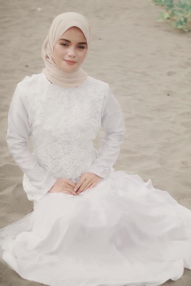 Beautiful islamic female model wearing hijab fashion, a modern wedding dress for muslim woman sitting in the sand and beach. Portrait an asian girl model using hijab having fun at the beach with trees photo