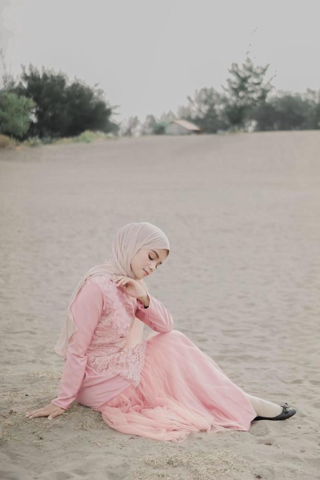 hermosa modelo femenina islámica usando moda hijab, un vestido de novia moderno para mujer musulmana sentada en la arena y la playa. retrata a una modelo asiática usando hiyab divirtiéndose en la playa con árboles foto