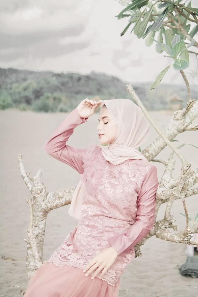 hermosa modelo femenina islámica usando moda hijab, un vestido de novia moderno para mujer musulmana sentada en la arena y la playa. retrata a una modelo asiática usando hiyab divirtiéndose en la playa con árboles foto