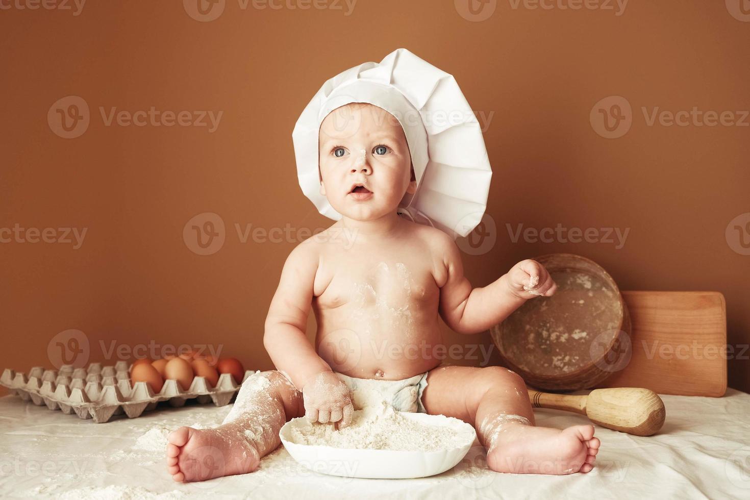 Little boy baker in a chef's hat sitting on the table playing with flour on a brown background with a wooden rolling pin, a round rustic sieve and eggs. Copy, empty space for text photo