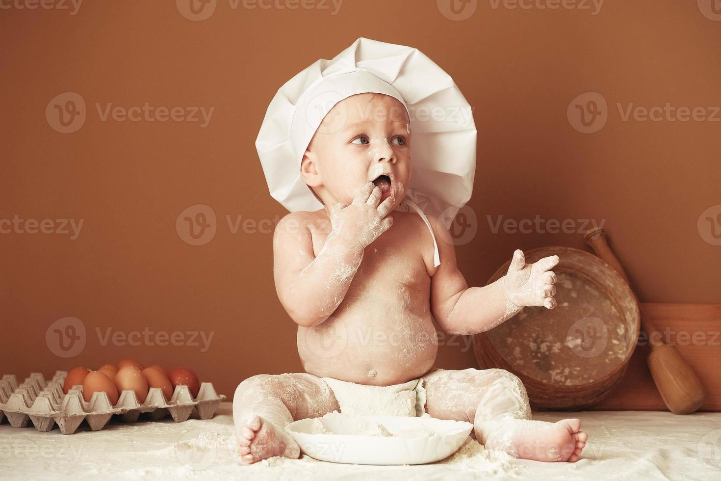 Little boy baker in a chef's hat sitting on the table playing with flour on a brown background with a wooden rolling pin, a round rustic sieve and eggs. Copy, empty space for text photo