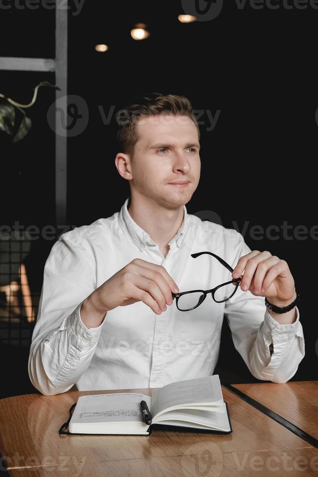 Young man wearing white shirt and eye glasses sitting at the table holding pen takes notes to his agenda on a dark background in office. Copy, empty space for text photo