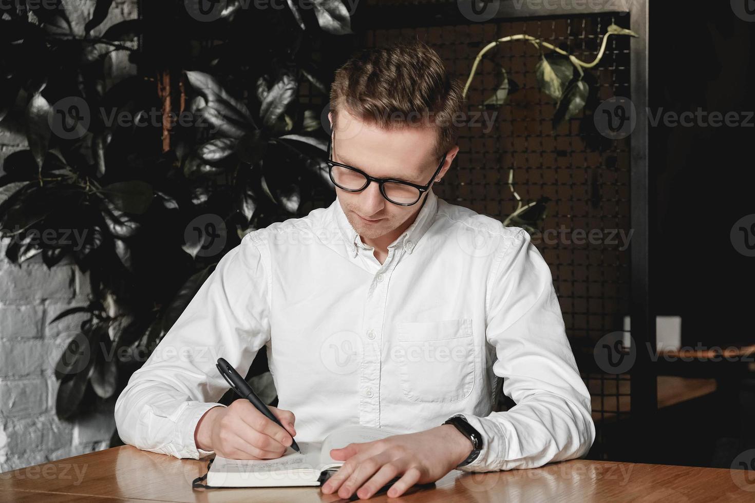 Serious young man wearing white shirt and eye glasses sitting at the table holding pen takes notes to his agenda on a dark background in office. Copy, empty space for text photo