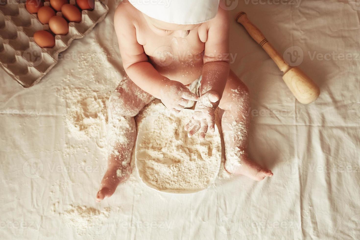 Little boy baker in a chef's hat sitting on the table playing with flour on a brown background with a wooden rolling pin, a round rustic sieve and eggs. Top view. Copy, empty space for text photo