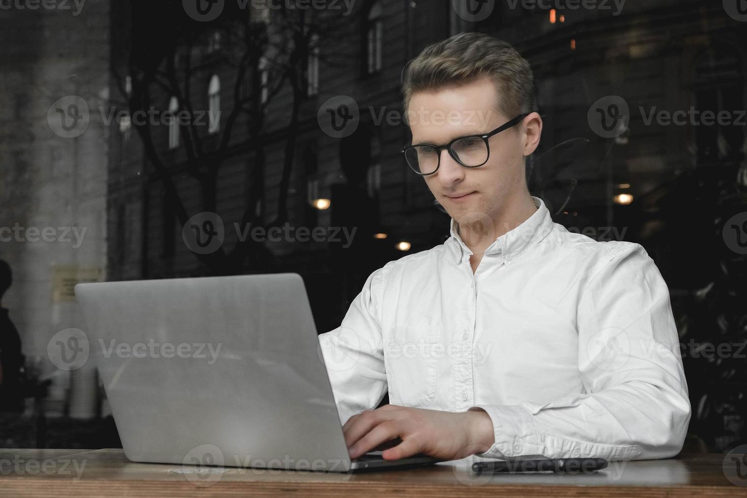 Young man wearing white shirt and eye glasses sitting with laptop and working in cafe. Freelance concept. Copy, empty space for text photo