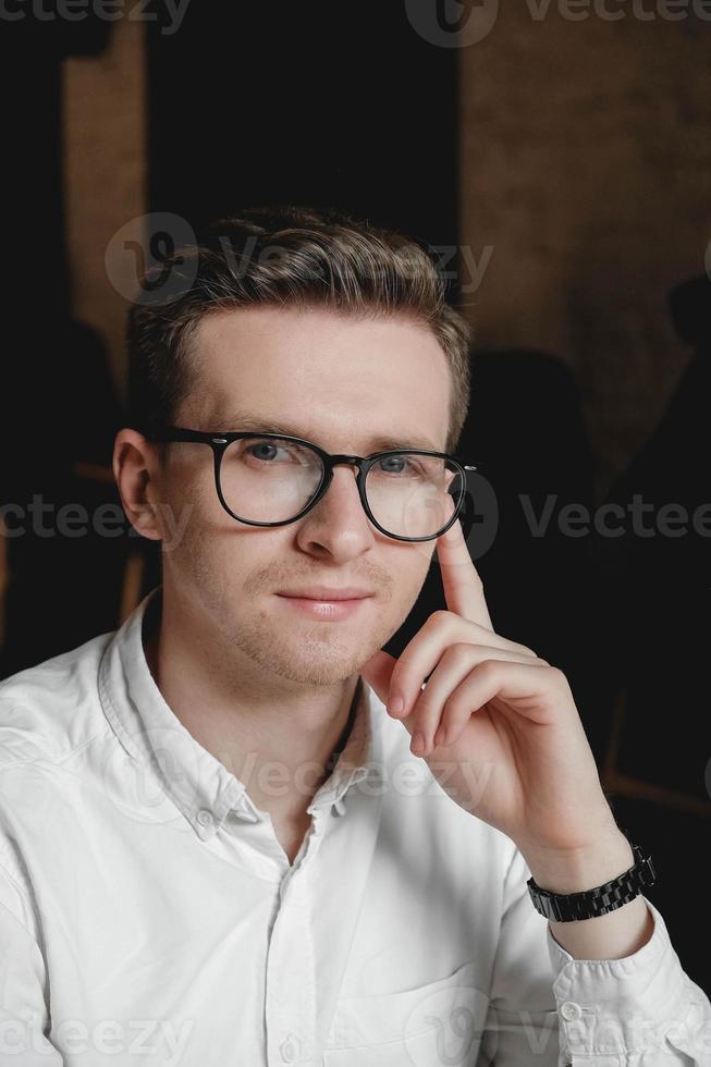 Portrait of a young man in glasses and a white shirt on a dark background. Copy, empty space for text photo