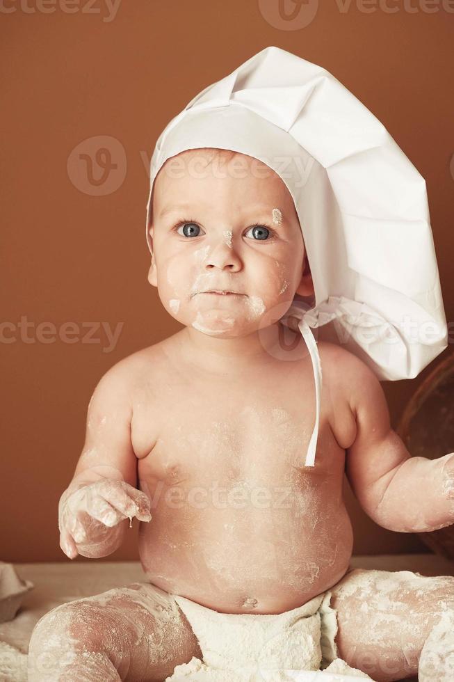 Little boy baker in a chef's hat sitting on the table playing with flour on a brown background with a wooden rolling pin, a round rustic sieve and eggs. Copy, empty space for text photo