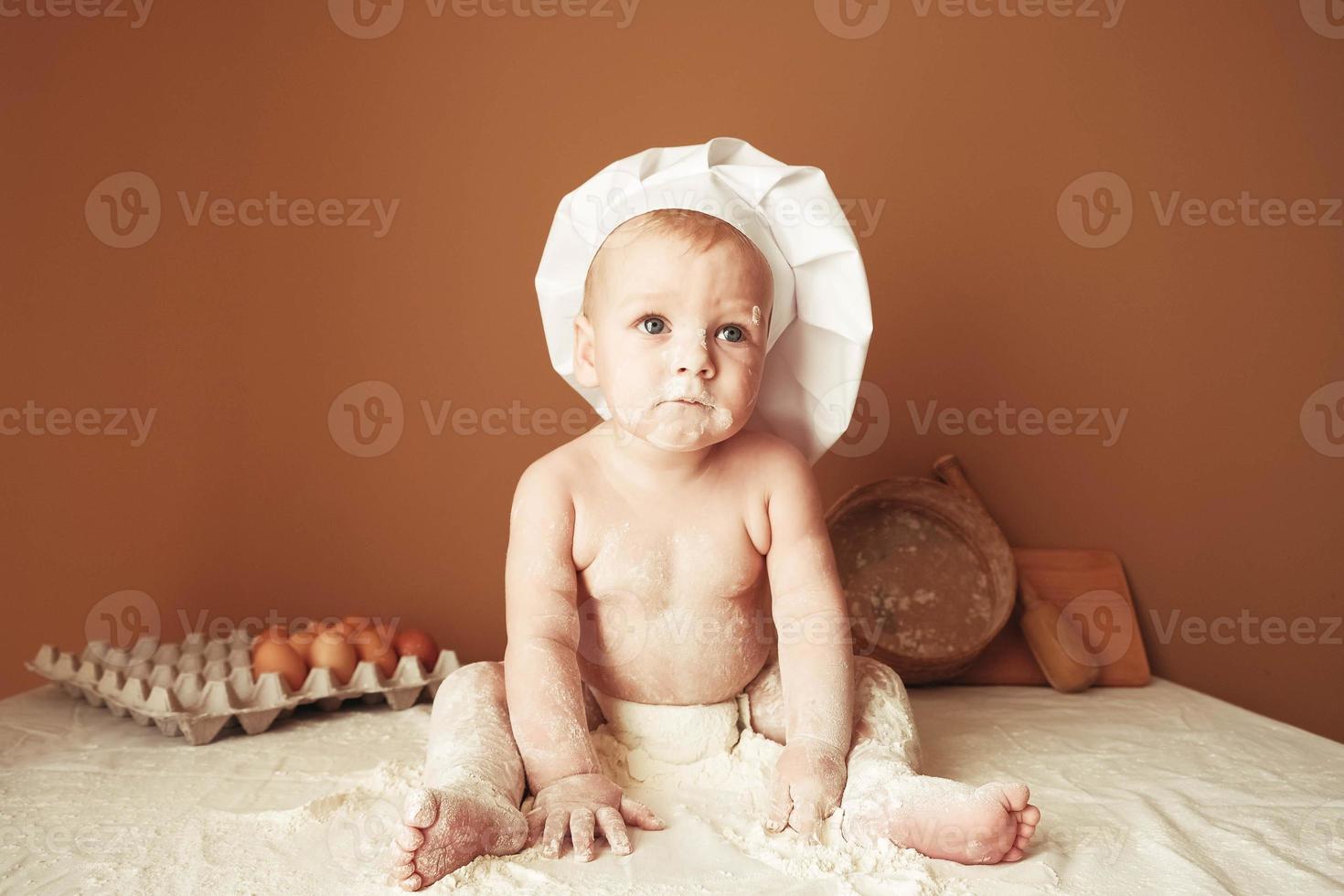 Little boy baker in a chef's hat sitting on the table playing with flour on a brown background with a wooden rolling pin, a round rustic sieve and eggs. Copy, empty space for text photo