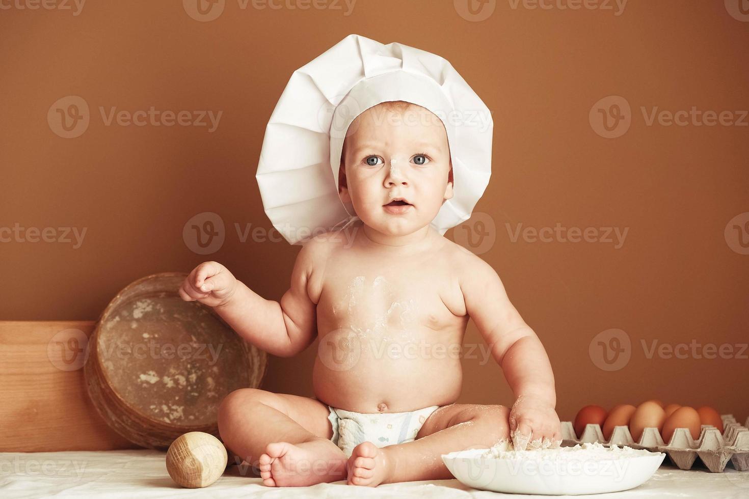 Little boy baker in a chef's hat sitting on the table playing with flour on a brown background with a wooden rolling pin, a round rustic sieve and eggs. Copy, empty space for text photo