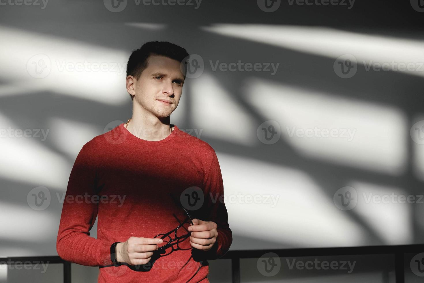 Portrait of young man holding eyeglasses with hands in a red sweater on a white background with shadows from the light photo