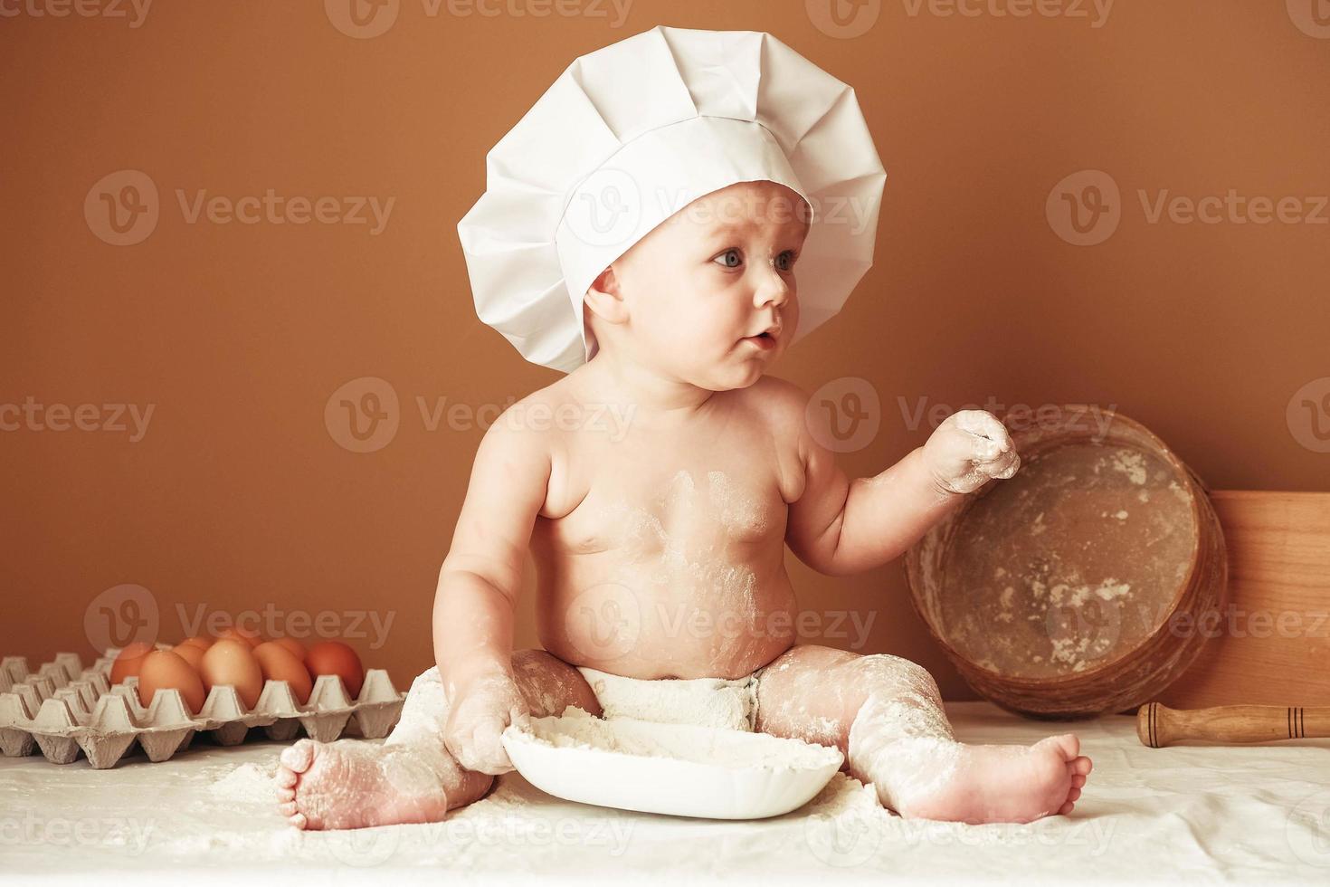 Little boy baker in a chef's hat sitting on the table playing with flour on a brown background with a wooden rolling pin, a round rustic sieve and eggs. Copy, empty space for text photo