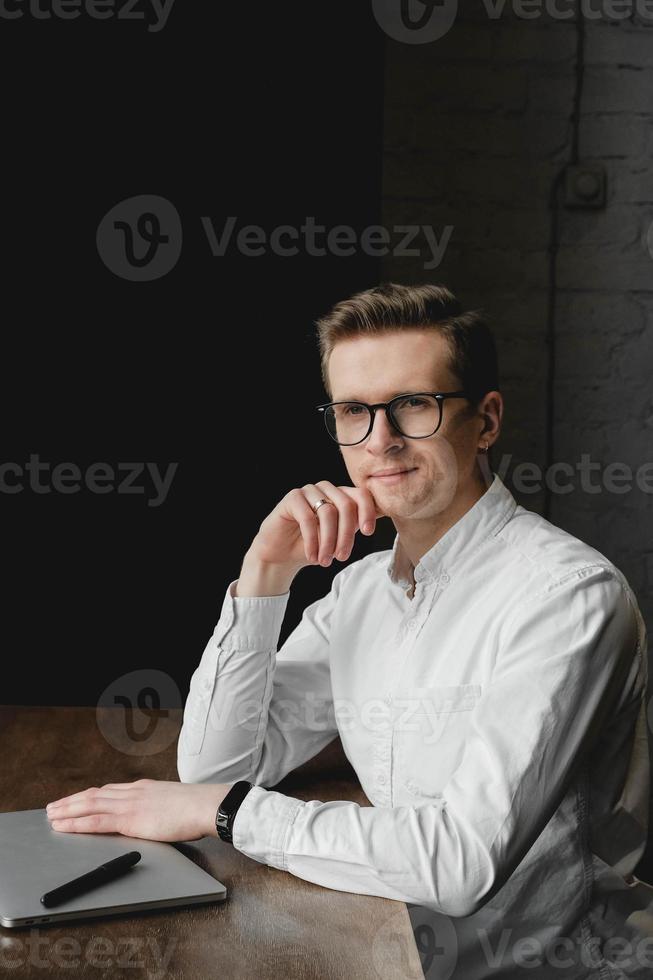 Portrait young man wearing white shirt and eye glasses sitting with laptop and working in cafe. Freelance concept. Copy, empty space for text photo