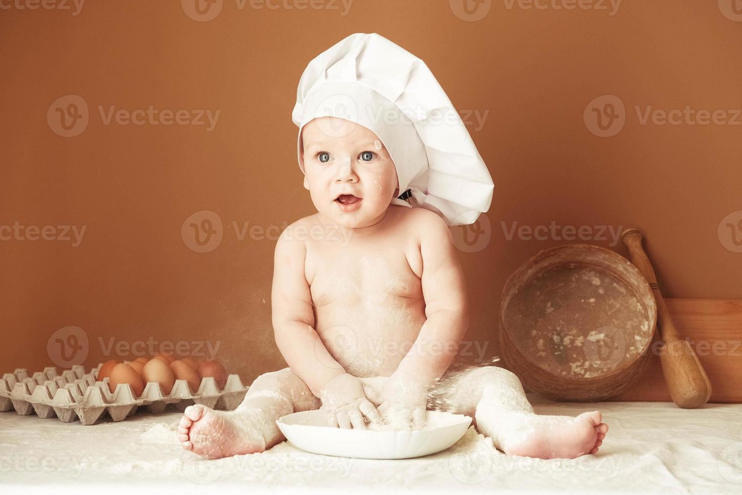 Little boy baker in a chef's hat sitting on the table playing with flour on a brown background with a wooden rolling pin, a round rustic sieve and eggs. Copy, empty space for text photo