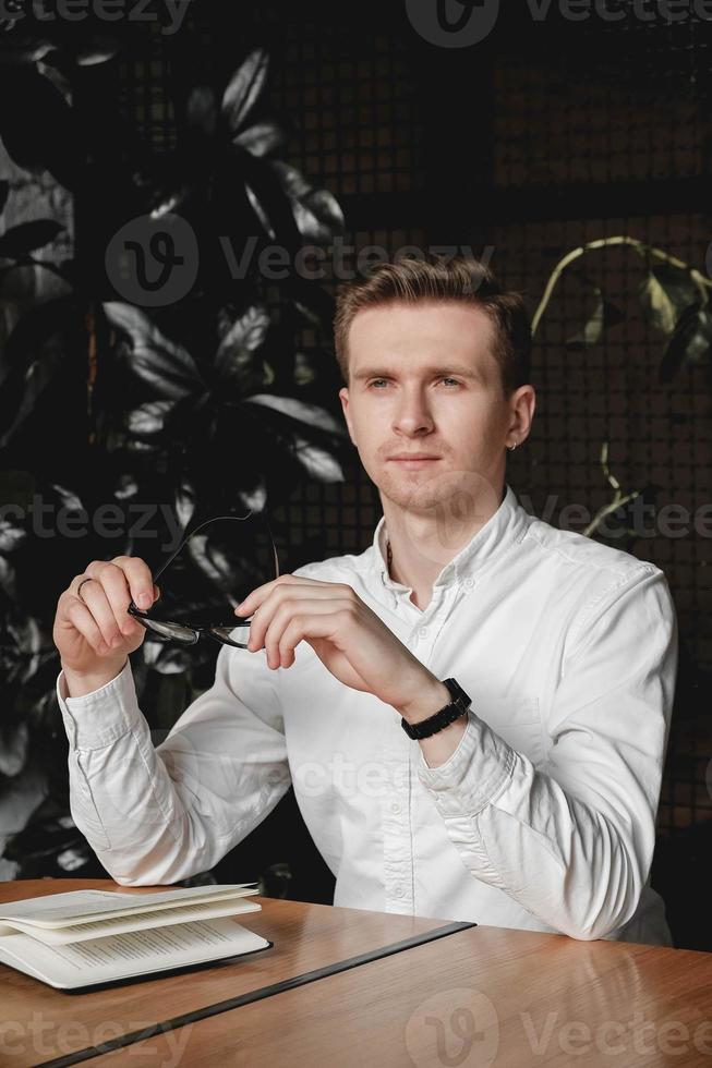 Young man wearing white shirt and eye glasses sitting at the table holding pen takes notes to his agenda on a dark background in office. Copy, empty space for text photo