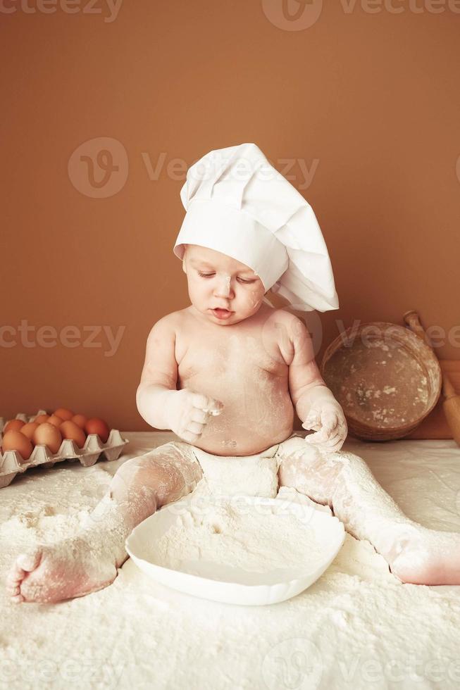 Little boy baker in a chef's hat sitting on the table playing with flour on a brown background with a wooden rolling pin, a round rustic sieve and eggs. Copy, empty space for text photo