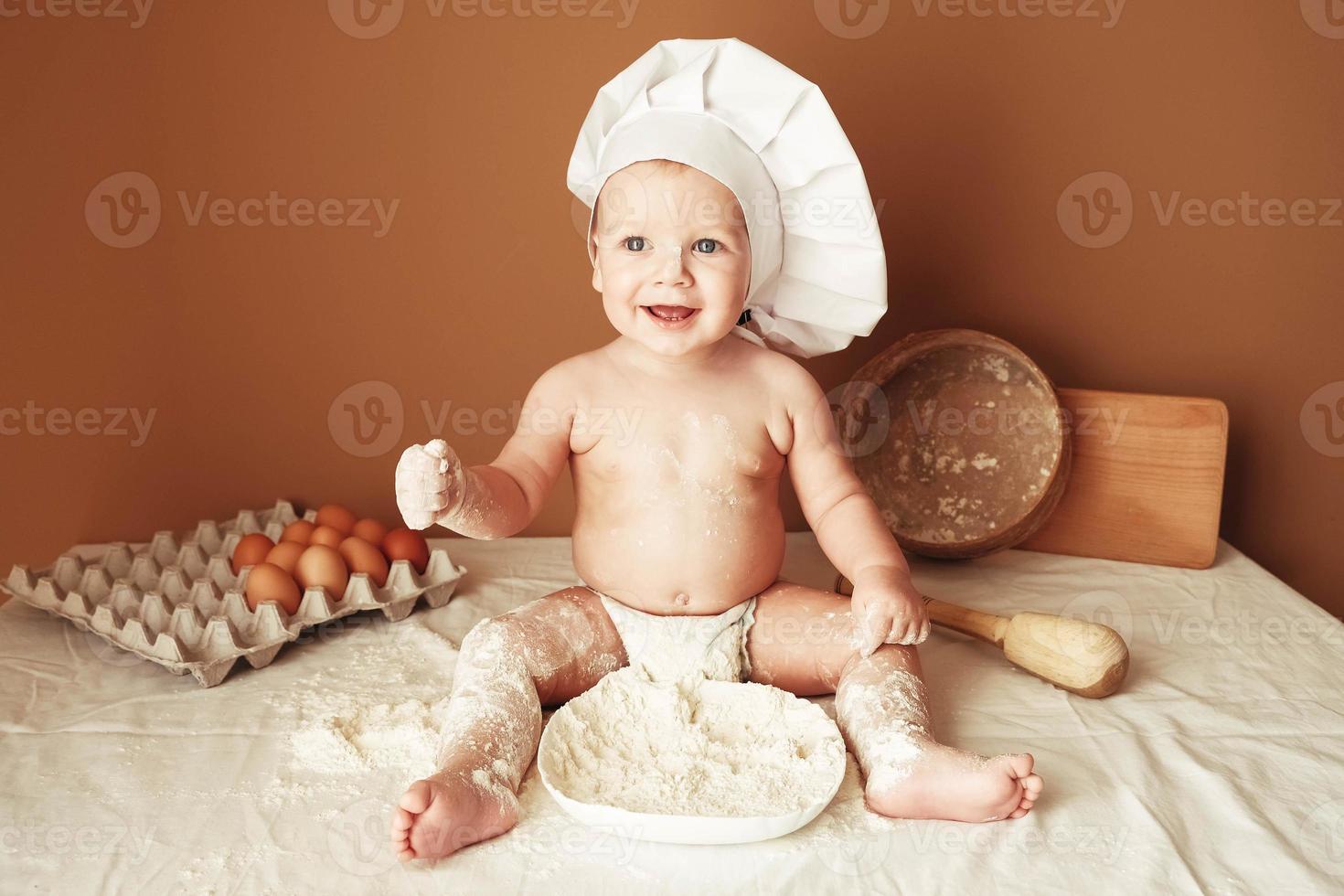 Little boy baker in a chef's hat sitting on the table playing with flour on a brown background with a wooden rolling pin, a round rustic sieve and eggs. Copy, empty space for text photo