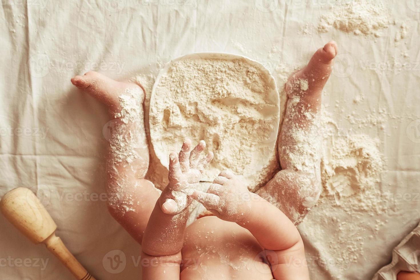 Little boy baker in a chef's hat sitting on the table playing with flour on a brown background with a wooden rolling pin, a round rustic sieve and eggs. Top view. Copy, empty space for text photo
