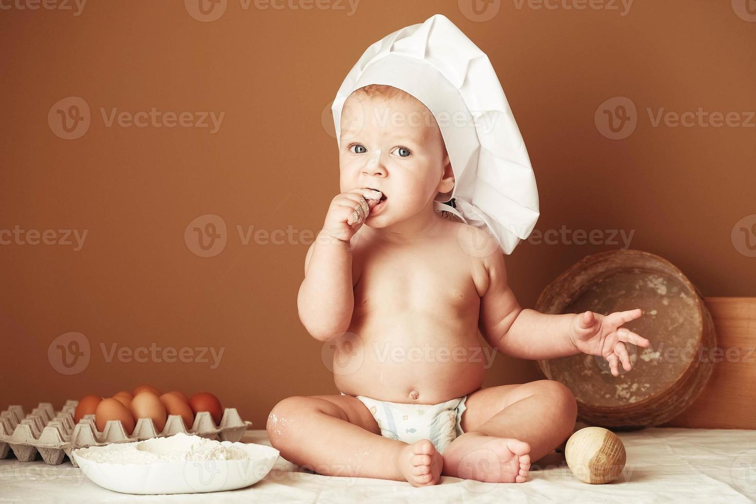 Little boy baker in a chef's hat sitting on the table playing with flour on a brown background with a wooden rolling pin, a round rustic sieve and eggs photo